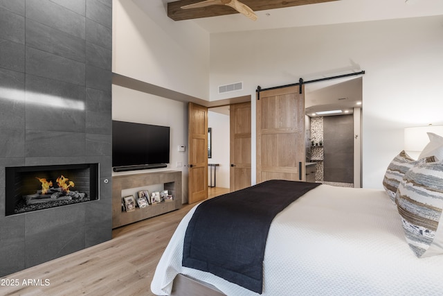 bedroom with a tile fireplace, beam ceiling, a high ceiling, light hardwood / wood-style floors, and a barn door