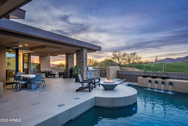 pool at dusk with a patio, a fire pit, area for grilling, pool water feature, and a mountain view