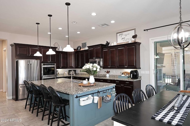 kitchen with light stone countertops, appliances with stainless steel finishes, dark brown cabinets, a kitchen island with sink, and decorative light fixtures