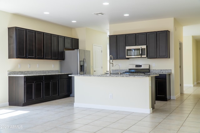 kitchen with a kitchen island with sink, light tile patterned floors, stainless steel appliances, light stone counters, and sink