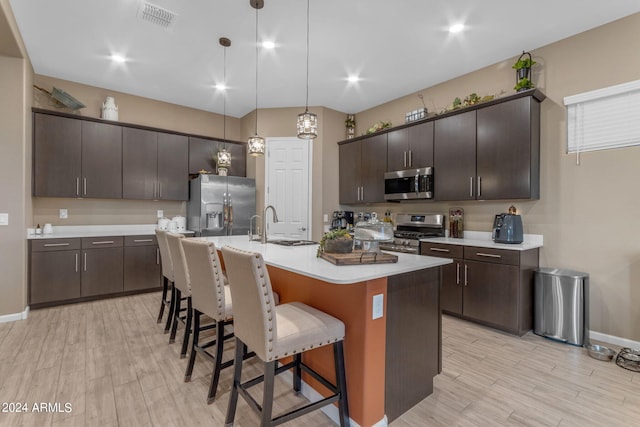 kitchen with an island with sink, hanging light fixtures, light hardwood / wood-style flooring, stainless steel appliances, and dark brown cabinetry