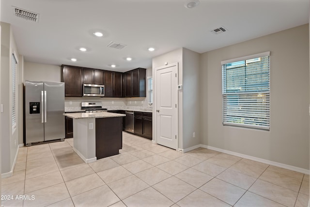 kitchen with appliances with stainless steel finishes, a center island, light tile patterned floors, light stone counters, and dark brown cabinets