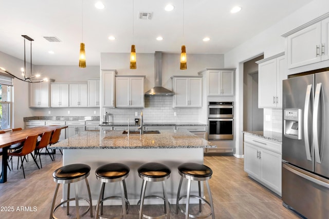 kitchen with visible vents, an island with sink, wall chimney exhaust hood, appliances with stainless steel finishes, and hanging light fixtures