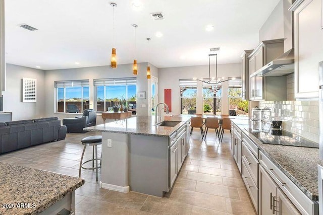 kitchen featuring pendant lighting, open floor plan, a sink, an island with sink, and wall chimney exhaust hood