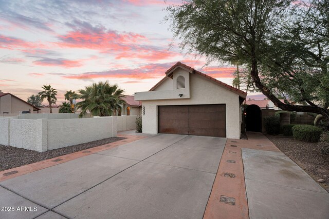 property exterior at dusk featuring an attached garage, concrete driveway, fence, stucco siding, and a tiled roof