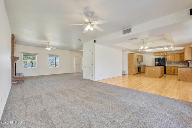 unfurnished living room featuring a brick fireplace, a tray ceiling, light colored carpet, and ceiling fan