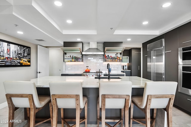 kitchen featuring a tray ceiling, wall chimney exhaust hood, built in refrigerator, and a kitchen breakfast bar