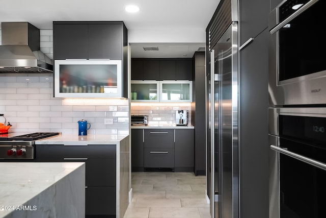 kitchen featuring decorative backsplash, wall chimney exhaust hood, light stone countertops, light tile patterned floors, and appliances with stainless steel finishes