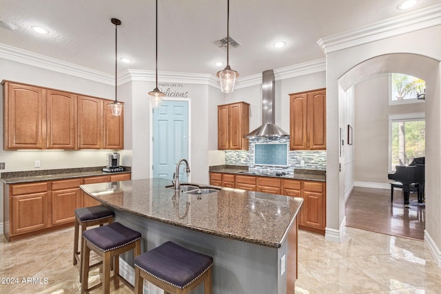 kitchen with sink, hanging light fixtures, ornamental molding, an island with sink, and wall chimney range hood