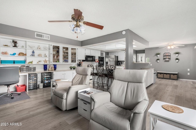 living room with ceiling fan, a textured ceiling, wine cooler, and light wood-type flooring