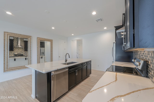 kitchen with wall chimney range hood, sink, an island with sink, appliances with stainless steel finishes, and tasteful backsplash