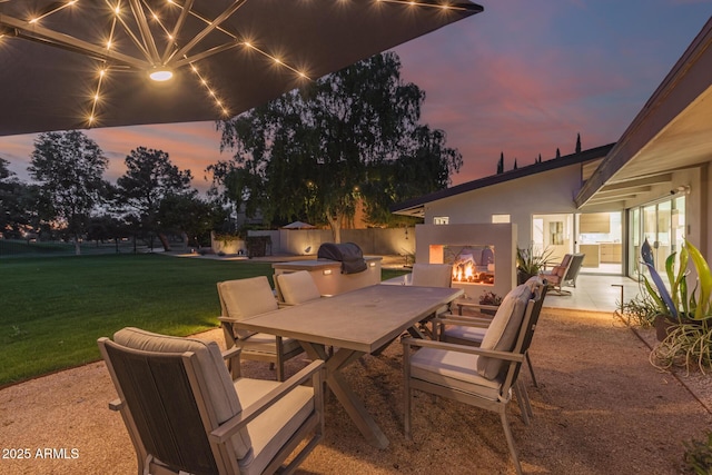 patio terrace at dusk with outdoor dining space, a multi sided fireplace, and a yard