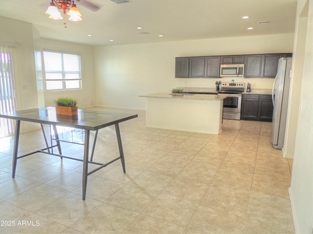 kitchen with light tile patterned flooring, dark brown cabinets, an island with sink, stainless steel appliances, and light stone countertops