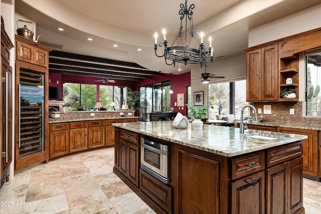 kitchen featuring sink, backsplash, light stone counters, decorative light fixtures, and an island with sink