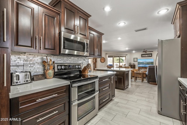 kitchen featuring kitchen peninsula, decorative backsplash, ceiling fan, dark brown cabinetry, and stainless steel appliances