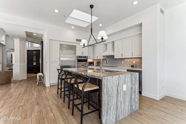 kitchen featuring built in refrigerator, decorative light fixtures, a center island with sink, stone counters, and white cabinets