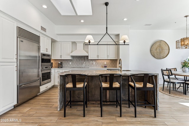 kitchen featuring premium range hood, appliances with stainless steel finishes, an island with sink, and pendant lighting