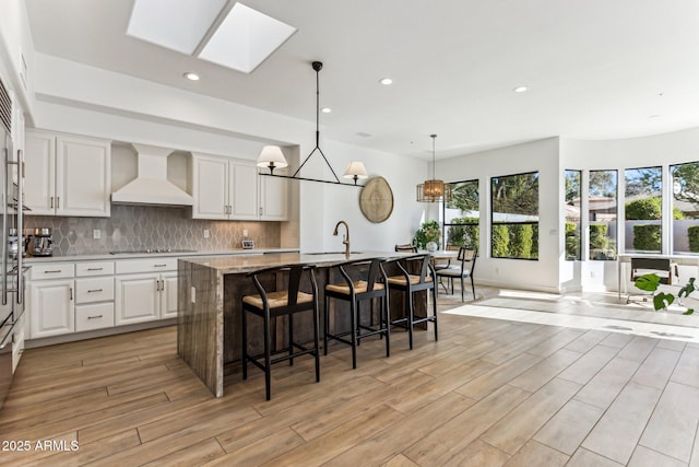 kitchen featuring sink, hanging light fixtures, white cabinets, a center island with sink, and custom exhaust hood