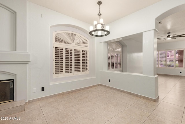 unfurnished dining area with arched walkways, ceiling fan with notable chandelier, baseboards, tile patterned floors, and a glass covered fireplace