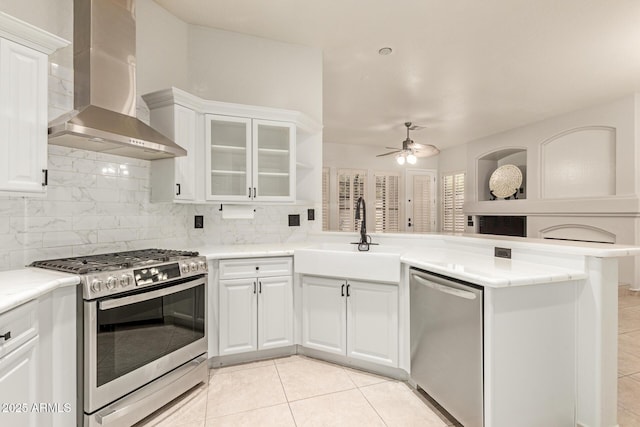 kitchen featuring decorative backsplash, appliances with stainless steel finishes, a peninsula, wall chimney range hood, and a sink