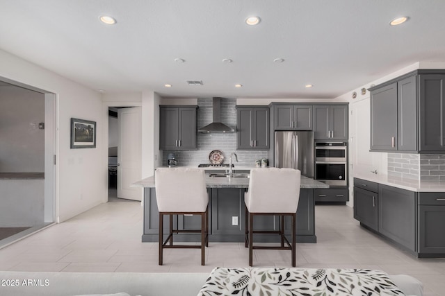 kitchen with wall chimney range hood, gray cabinets, appliances with stainless steel finishes, backsplash, and a center island with sink