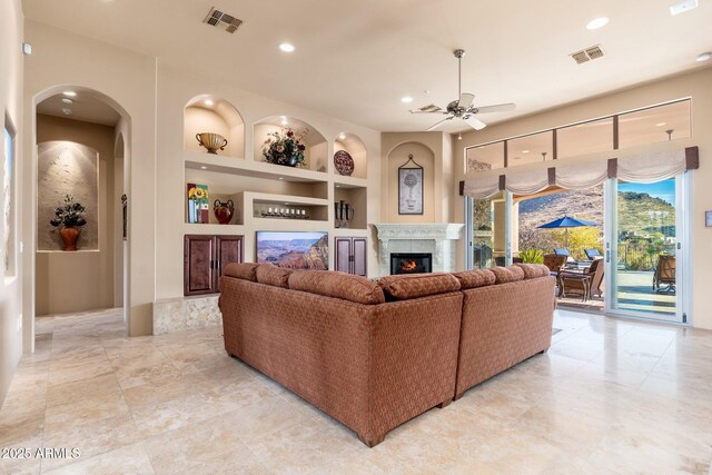 living area featuring a warm lit fireplace, ceiling fan, built in shelves, and visible vents