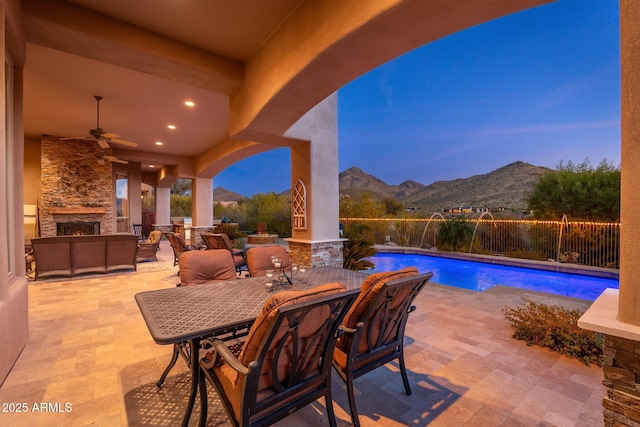 patio terrace at dusk with a mountain view, a ceiling fan, fence, an outdoor stone fireplace, and outdoor dining area