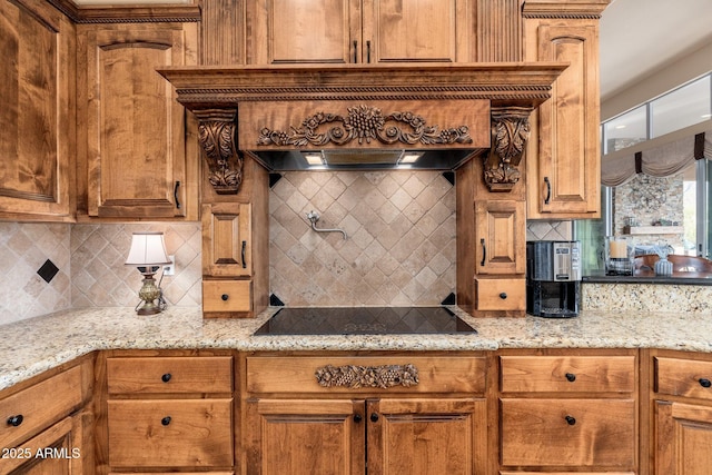 kitchen with custom exhaust hood, light stone counters, black electric stovetop, brown cabinets, and backsplash