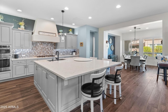 kitchen featuring dark wood-style floors, backsplash, a sink, an island with sink, and premium range hood