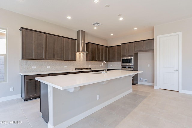 kitchen with dark brown cabinetry, a center island with sink, light tile patterned floors, stainless steel appliances, and wall chimney range hood