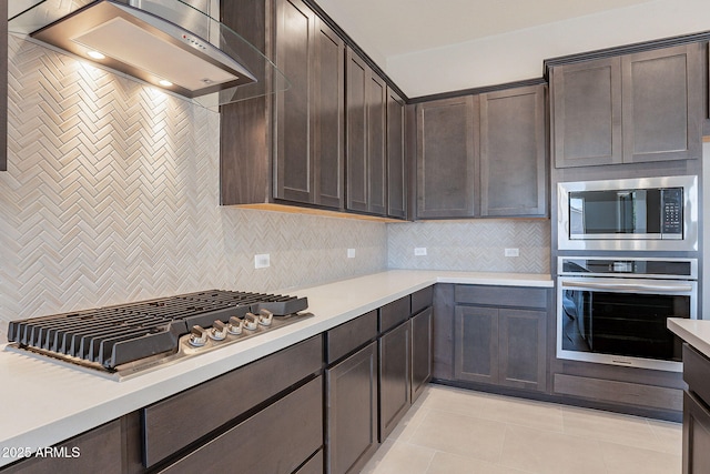 kitchen featuring wall chimney range hood, light tile patterned floors, appliances with stainless steel finishes, dark brown cabinets, and decorative backsplash