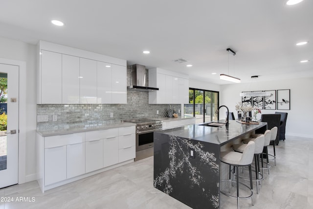 kitchen with stainless steel stove, decorative light fixtures, a center island with sink, wall chimney exhaust hood, and white cabinets