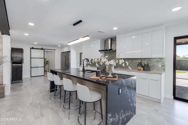 kitchen with white cabinetry, wall chimney exhaust hood, a barn door, and a kitchen island with sink