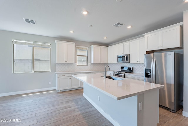 kitchen featuring appliances with stainless steel finishes, light wood-style floors, decorative backsplash, white cabinetry, and a kitchen island with sink