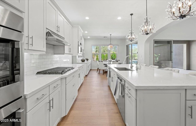 kitchen with tasteful backsplash, stainless steel appliances, under cabinet range hood, a sink, and a notable chandelier