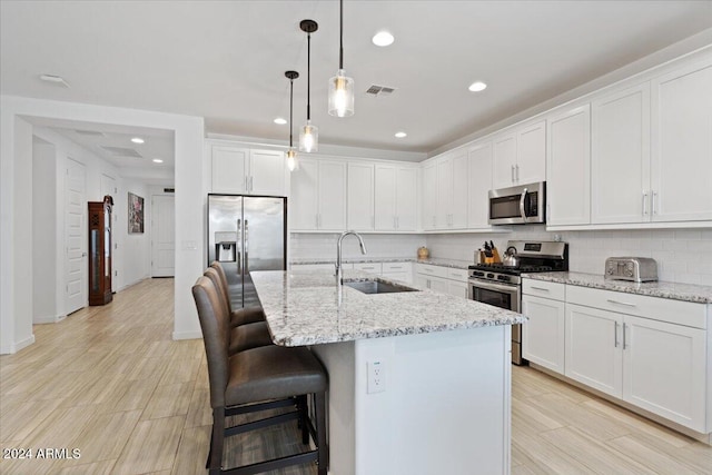 kitchen with light stone counters, stainless steel appliances, white cabinetry, decorative light fixtures, and an island with sink