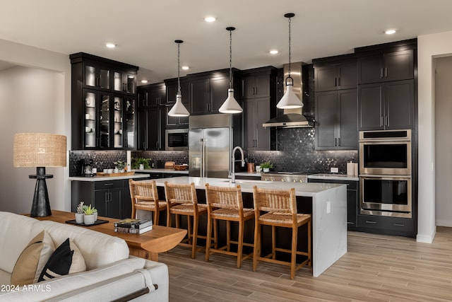 kitchen with built in appliances, decorative light fixtures, a center island with sink, and wall chimney range hood