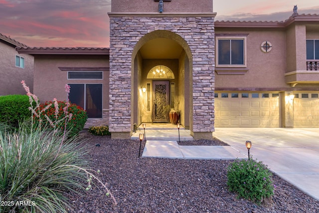 exterior entry at dusk featuring an attached garage, a tile roof, concrete driveway, and stucco siding