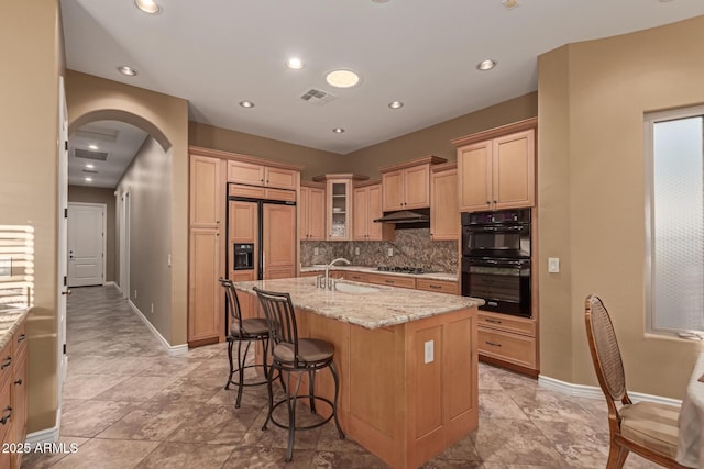 kitchen featuring sink, tasteful backsplash, light stone countertops, an island with sink, and black double oven