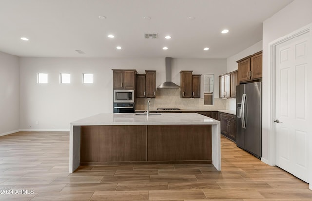 kitchen with tasteful backsplash, stainless steel appliances, a kitchen island with sink, sink, and wall chimney range hood