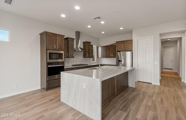 kitchen with a kitchen island with sink, sink, wall chimney exhaust hood, light stone countertops, and stainless steel appliances