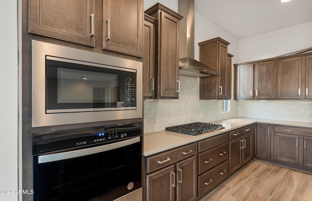 kitchen featuring dark brown cabinetry, wall chimney range hood, light hardwood / wood-style flooring, backsplash, and appliances with stainless steel finishes