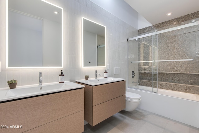 bathroom featuring two vanities, bath / shower combo with glass door, a sink, tile walls, and toilet