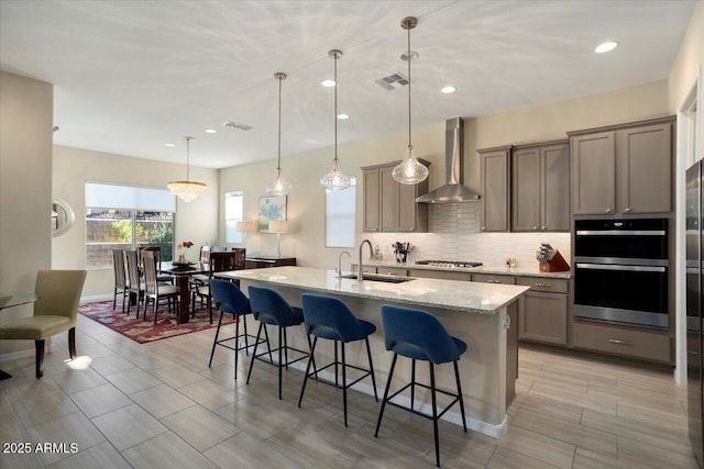 kitchen featuring light stone countertops, appliances with stainless steel finishes, wall chimney exhaust hood, sink, and pendant lighting