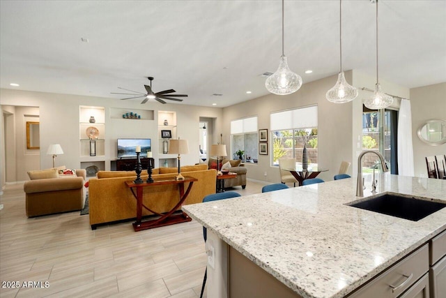 kitchen featuring light stone countertops, ceiling fan, a kitchen island with sink, sink, and hanging light fixtures