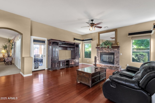 living area featuring ceiling fan, arched walkways, a stone fireplace, dark wood-style floors, and baseboards