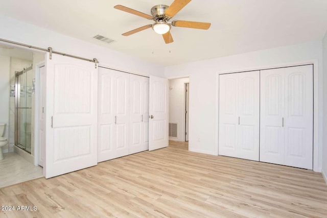 unfurnished bedroom featuring ceiling fan, ensuite bathroom, light hardwood / wood-style flooring, and a barn door