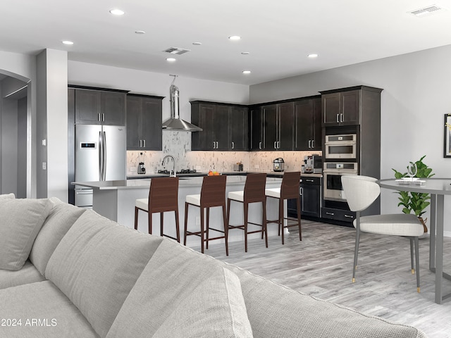 kitchen featuring appliances with stainless steel finishes, light wood-type flooring, a center island with sink, tasteful backsplash, and wall chimney range hood