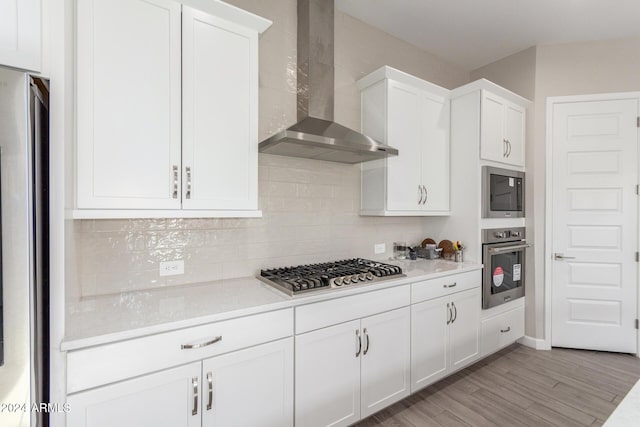 kitchen with white cabinets, wall chimney range hood, light hardwood / wood-style flooring, appliances with stainless steel finishes, and tasteful backsplash