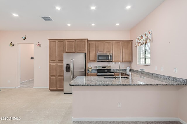 kitchen with appliances with stainless steel finishes, a peninsula, light stone counters, brown cabinetry, and recessed lighting